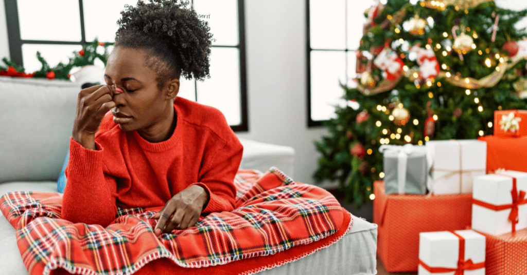 Black woman in a red sweater sitting near Christmas tree pinching her nose