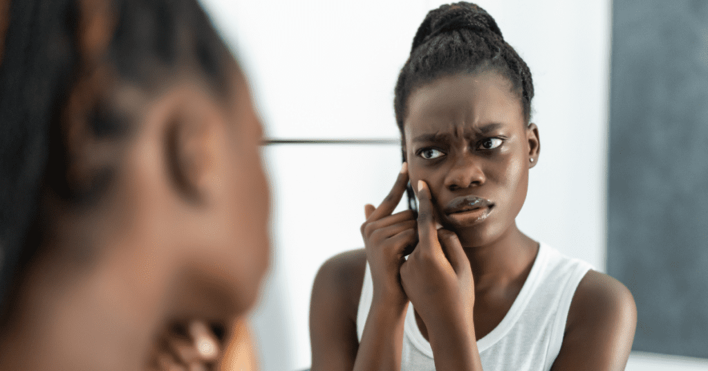 Young Black woman picking at skin in the mirror