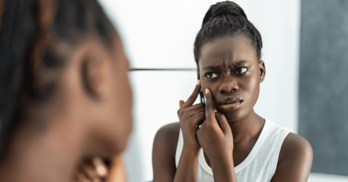 Young Black woman picking at skin in the mirror