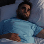 Man with beard laying in bed with a troubled look on his face.