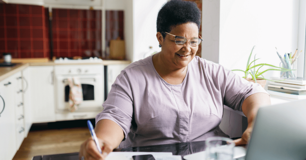 Black woman in purple shirt and glasses in front of a laptop taking notes with a pen