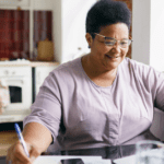 Black woman in purple shirt and glasses in front of a laptop taking notes with a pen
