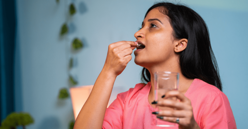 South asian woman in a pink shirt holding a glass of water in her left hand and putting a statin pill in her mouth with the right hand