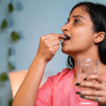 South asian woman in a pink shirt holding a glass of water in her left hand and putting a statin pill in her mouth with the right hand