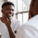 5 Skin Conditions Men Are More Susceptible To 5 Black man looking in the mirror and applying a skincare product to his face