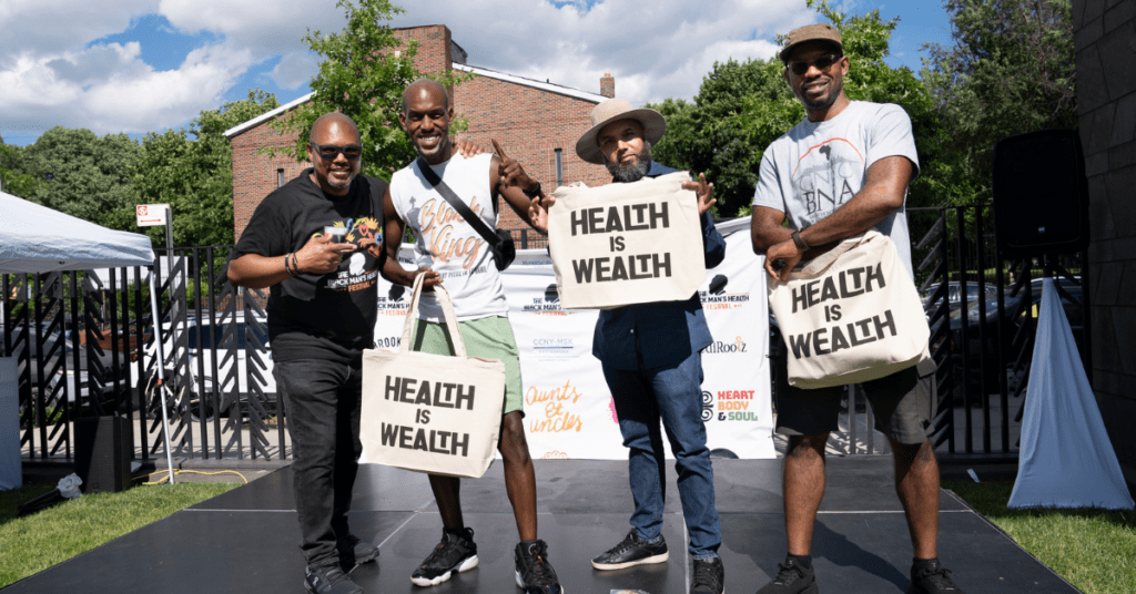 Cofounder of Heart, Body and Soul, Christopher Williams, on a stage with Black men holding up “Health is Wealth” tote bags