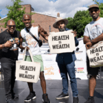 Cofounder of Heart, Body and Soul, Christopher Williams, on a stage with Black men holding up “Health is Wealth” tote bags