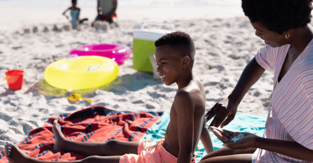 African american mother applying sunscreen on shirtless son's back while sitting on beach.