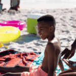 Is Your "Waterproof" Sunscreen Lying to You? 4 African american mother applying sunscreen on shirtless son's back while sitting on beach.
