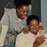 Elderly African American woman sitting on a couch holding her phone in one hand, and a walking cane in another. She is accompanied by her daughter who is standing above her shoulder, smiling.