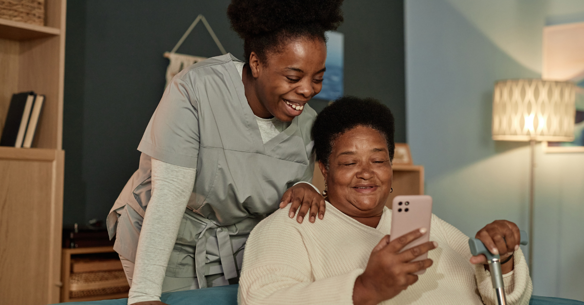 Elderly African American woman sitting on a couch holding her phone in one hand, and a walking cane in another. She is accompanied by her daughter who is standing above her shoulder, smiling.
