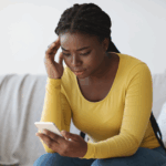 Black woman on couch, rubbing the temples of her head while she reads her phone