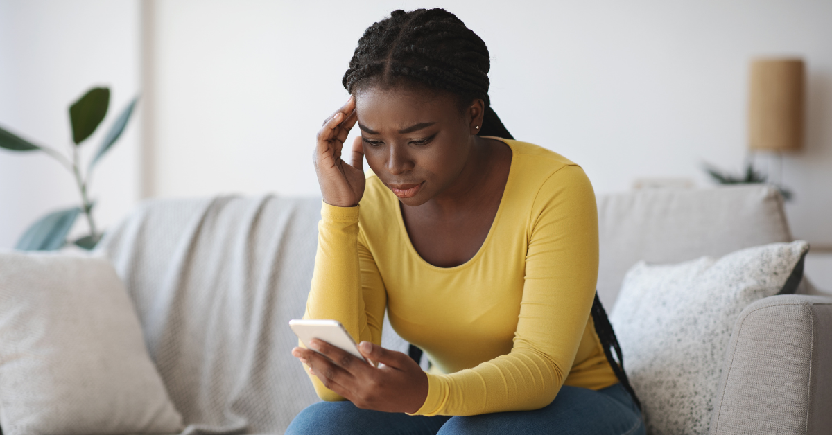 Black woman on couch, rubbing the temples of her head while she reads her phone