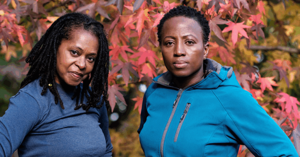 Two mature Black women in fitness attire standing in front of an autumn tree, looking directly at the camera