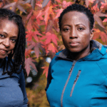 Two mature Black women in fitness attire standing in front of an autumn tree, looking directly at the camera