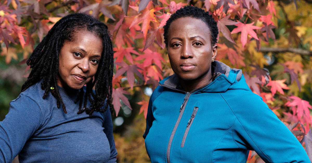Two mature Black women in fitness attire standing in front of an autumn tree, looking directly at the camera
