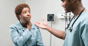 Black woman in a patient room holding her neck and explaining her symptoms to a South asian doctor
