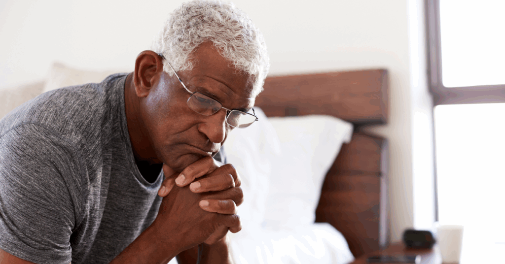 Close-up of a tired elderly Black man sitting on the edge of his bed, resting his chin on his hands