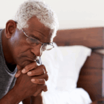 Close-up of a tired elderly Black man sitting on the edge of his bed, resting his chin on his hands