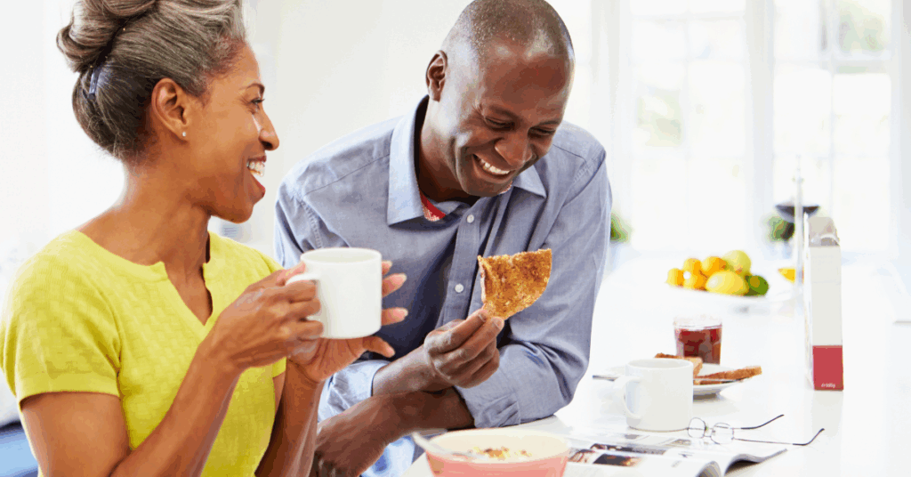 Middle-aged Black couple eating breakfast. The woman is holding a cup while the man is eating toast.