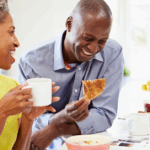 Middle-aged Black couple eating breakfast. The woman is holding a cup while the man is eating toast.