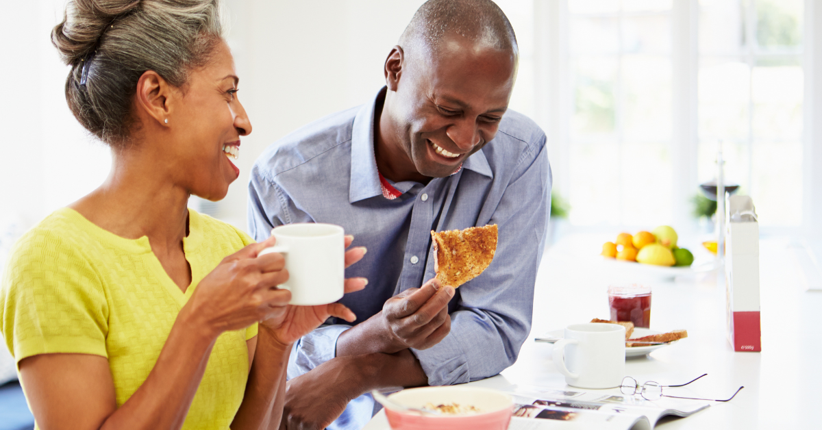 Middle-aged Black couple eating breakfast. The woman is holding a cup while the man is eating toast.