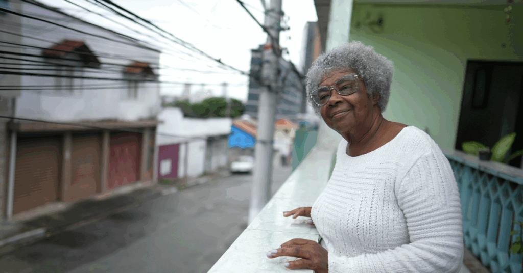 Elderly Black woman with glasses standing on her balcony, with a rural neighbourhood in the background.