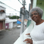 Elderly Black woman with glasses standing on her balcony, with a rural neighbourhood in the background.