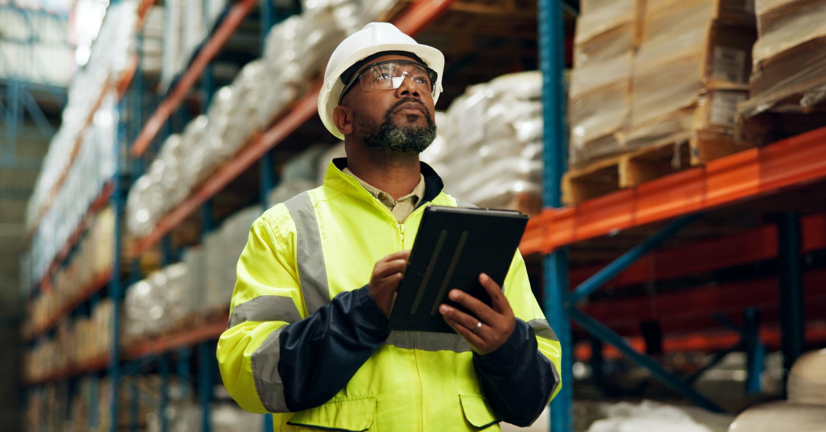 Black man in a hard hat, eye goggles, and yellow jacket, holding a tablet and looking up. There is a warehouse behind him, implying that he may be exposed to risk factors for lymphoma.