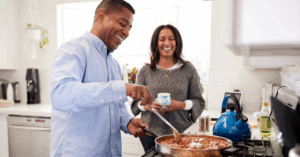 Millennial Black man cooking food, which looks like a meat stew, on the stove, while his partner stands beside him smiling with a cup in her hand
