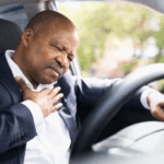 Middle-aged Black man wearing a suit in a car, clutching his heart with his right hand and the steering wheel with his left hand, indicating that he may have a type of heart disease.