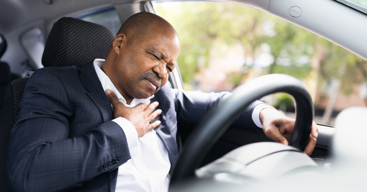 Middle-aged Black man wearing a suit in a car, clutching his heart with his right hand and the steering wheel with his left hand, indicating that he may have a type of heart disease.