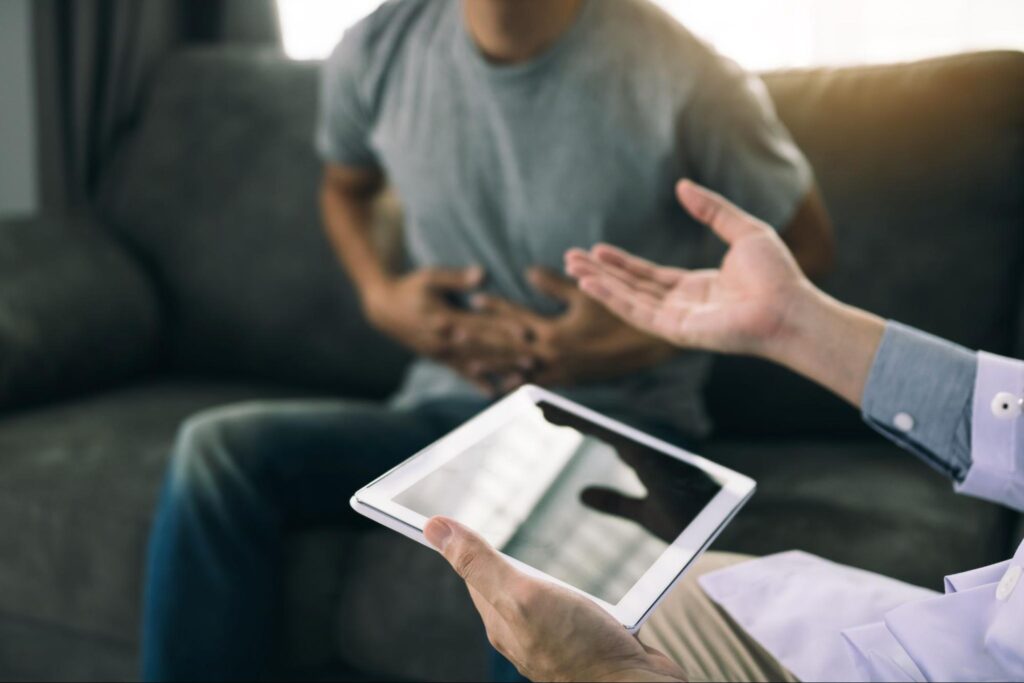 Close-up of man clutching his stomach due to ulcerative colitis, while sitting on the couch, consulting a healthcare professional