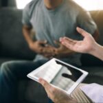 Close-up of man clutching his stomach due to ulcerative colitis, while sitting on the couch, consulting a healthcare professional