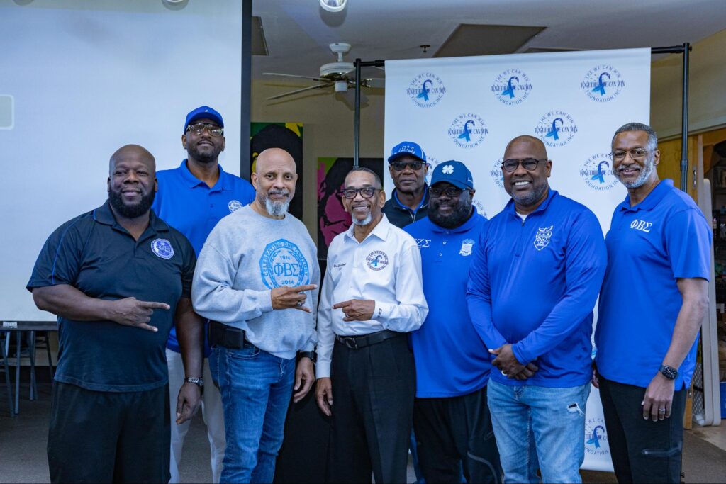 Dr. Edward Wright, founder of The We Can Win Foundation and a group of seven Black men, most wearing blue shirts, pose for a group photo indoors.