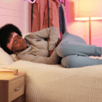 Young Black woman in a gray sweatshirt and blue joggers, laying in bed, crutching her stomach in pain; likely due to a UC flare-up