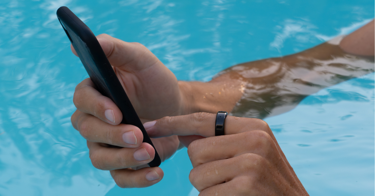 Close-up of a person’s hands in the swimming pool, wearing an oura ring and using their phone
