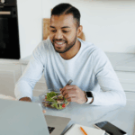 Smiling african american man eating fresh salad and using laptop near notebooks in kitchen, implying he’s using a monthly plan to lower his high blood pressure