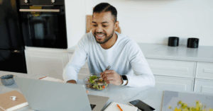 Smiling african american man eating fresh salad and using laptop near notebooks in kitchen, implying he’s using a monthly plan to lower his high blood pressure