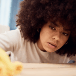 Close-up of young Black woman with an afro, wearing yellow gloves, and cleaning her shelf with a spray that contains forever chemicals (PFAS)