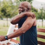Hidden Signs of Lung Cancer in Non-Smokers 5 Older Black man sitting on a bench in a park, clutching his left shoulder with his right hand, likely due to pain from lung cancer