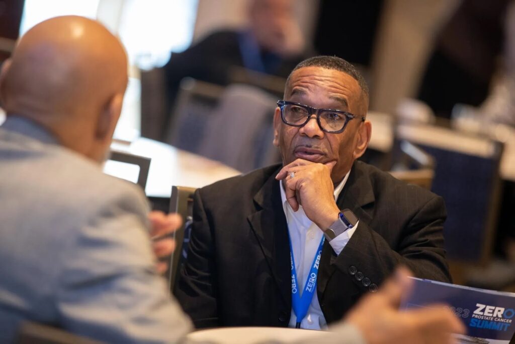 Dr. Edward Wright in a black blazer and glasses sits at a table, looking intently at another person out of frame to his left.