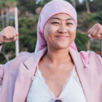 Young indigenous woman in a pink head scarf, indicating she might be living with breast cancer, smiling at the camera and flexing her biceps.