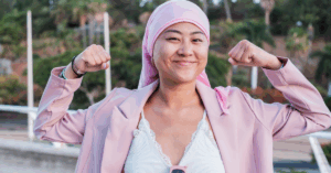 Young indigenous woman in a pink head scarf, indicating she might be living with breast cancer, smiling at the camera and flexing her biceps.