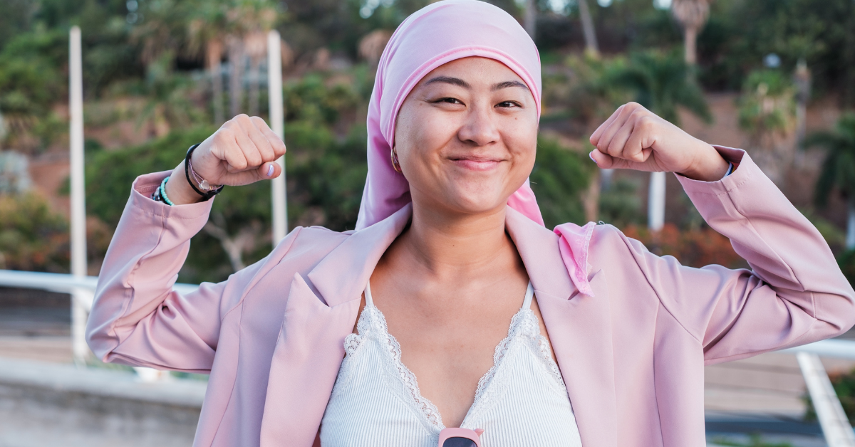Young indigenous woman in a pink head scarf, indicating she might be living with breast cancer, smiling at the camera and flexing her biceps.