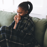 Young Black woman with allergies wrapped in blanket on couch sneezing into tissue, surrounded by pillows and used napkins, showing symptoms of cold or flu in cozy home setting.