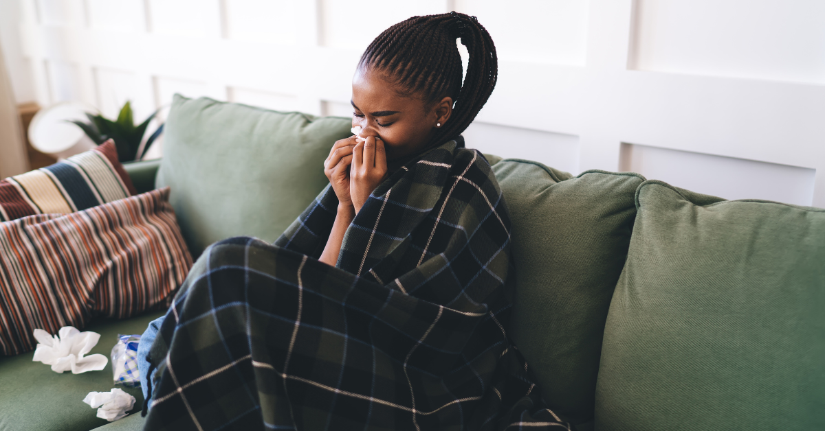 Young Black woman with allergies wrapped in blanket on couch sneezing into tissue, surrounded by pillows and used napkins, showing symptoms of cold or flu in cozy home setting.