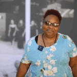 NOWINCLUDED community member, Janiqua, in a light blue floral dress and sunglasses with short reddish hair, smiling and talking about her journey with aortic stenosis.