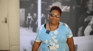 NOWINCLUDED community member, Janiqua, in a light blue floral dress and sunglasses with short reddish hair, smiling and talking about her journey with aortic stenosis.