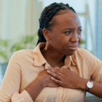 A young Black woman sitting on the couch looking distressed while putting both hands on her chest, likely due to an irregular heartbeat